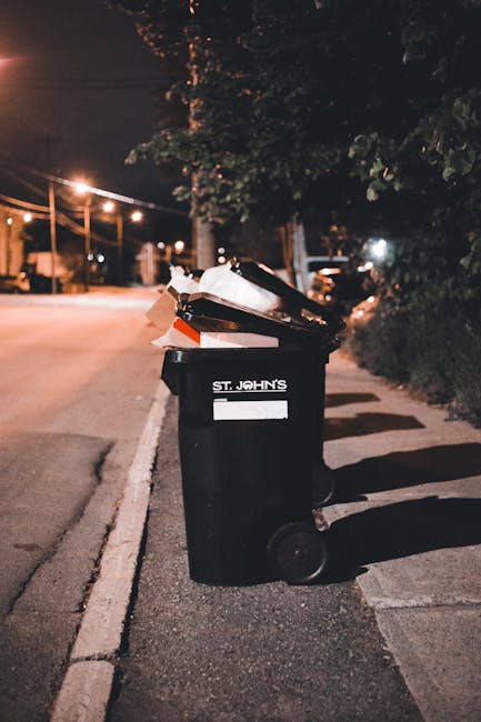 A row of large, rectangular outdoor waste bins made of durable plastic with hinged lids, positioned along a pavement beside a brick wall. The bins are primarily green and blue, with some red lids on t